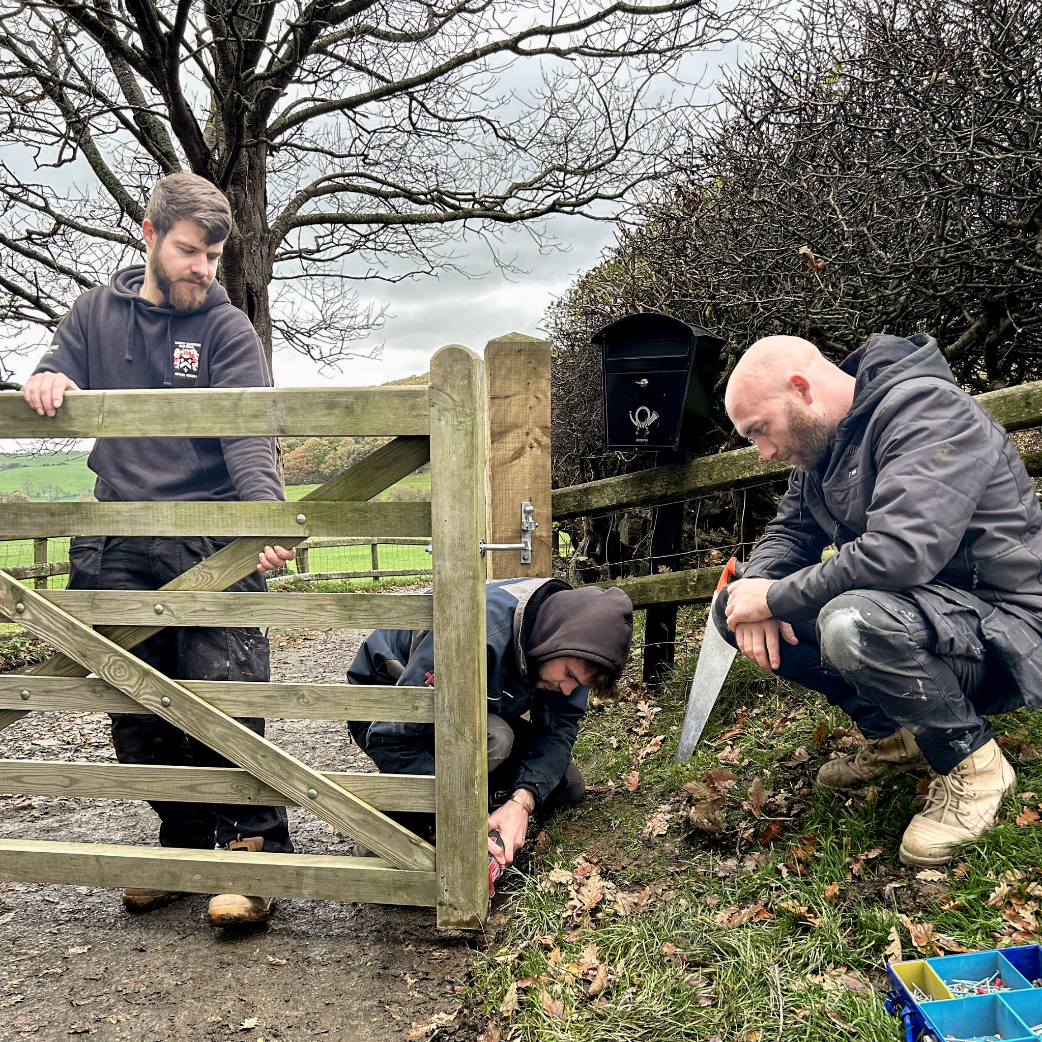 Home A group of three Sonny Vaughan Electrical electricians installing an electric gate outdoors. One electrician is holding up the gate while the other two are knelt on the ground cutting the bottom.
