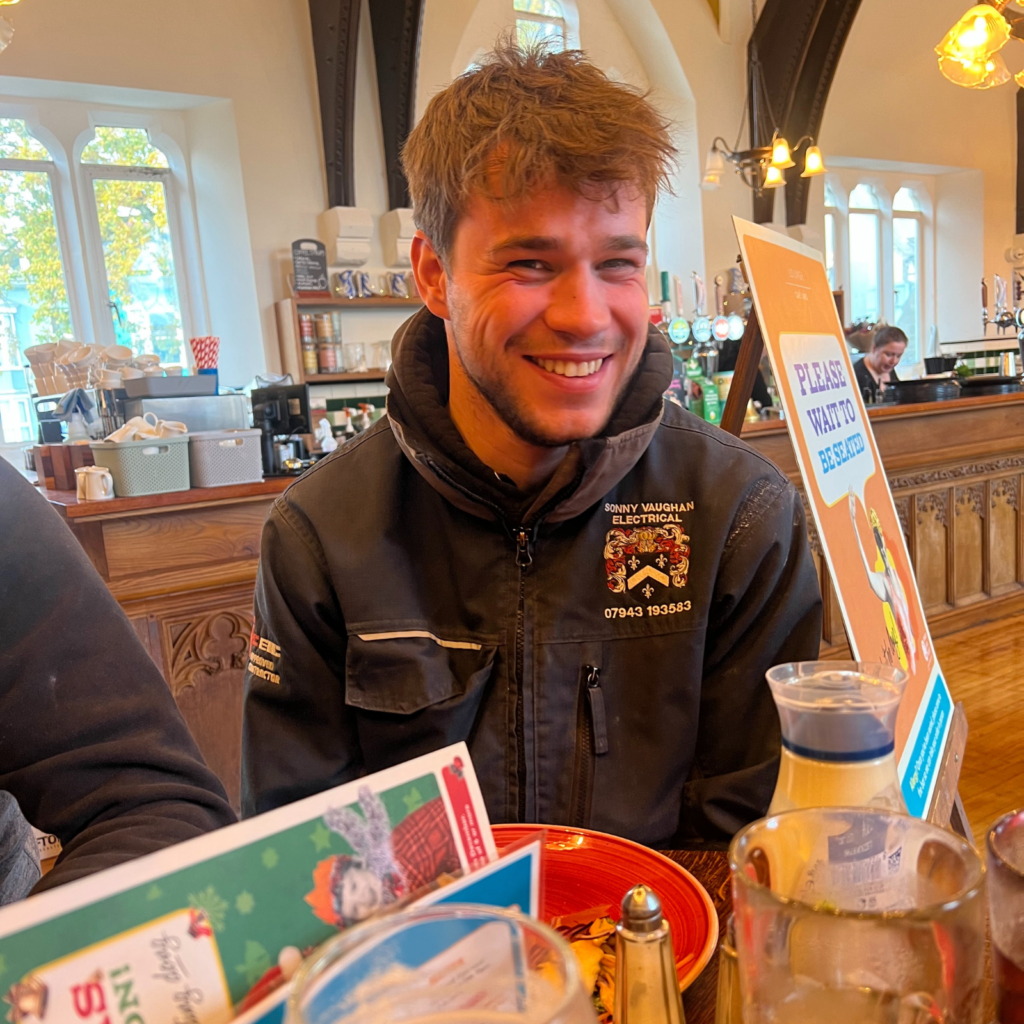 Smiling electrician wearing Sonny Vaughan Electrical uniform. The electrician is at a table in a cafe.