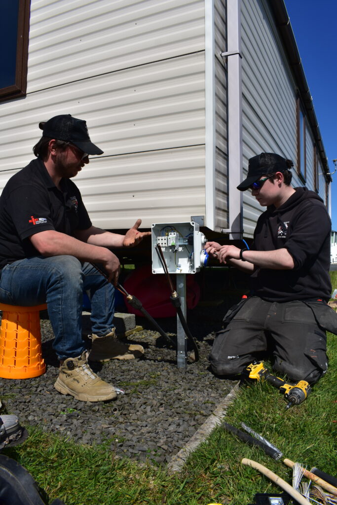 electricians at a caravan park installing a power box in front of a static caravan.