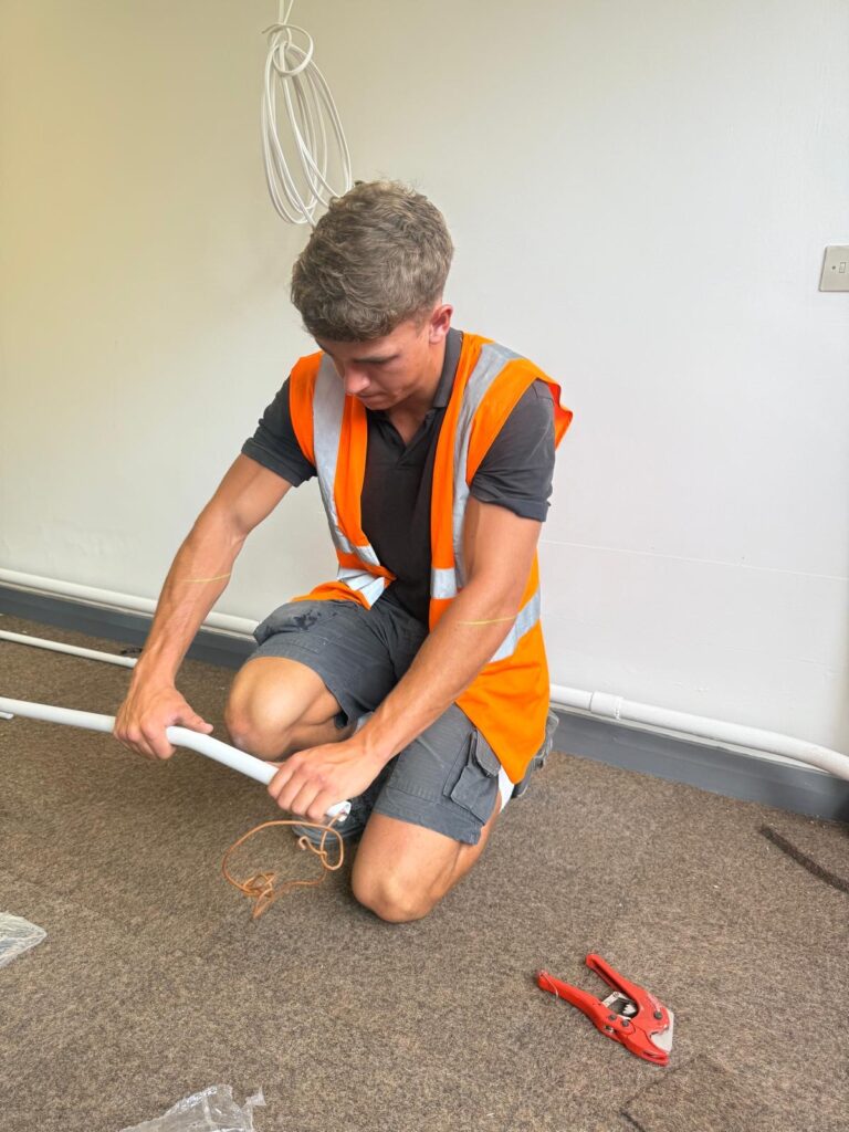 Electrician in orange high-vis, kneeling down while bending some piping.