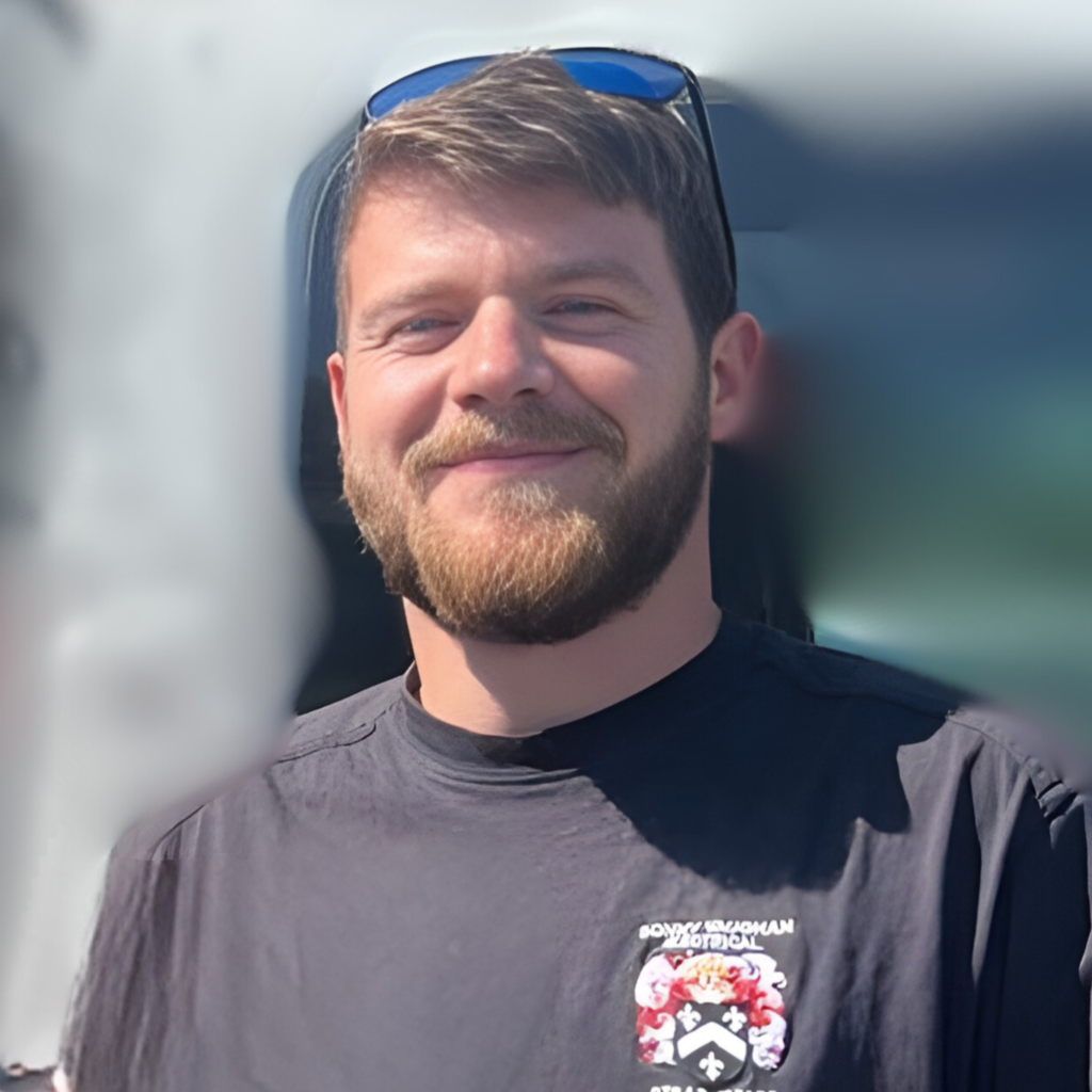 Headshot of electrician with short brown and blue glasses on top of head in black Sonny Vaughan Electrical t-shirt smiling at the camera. He has a beard and moustache.