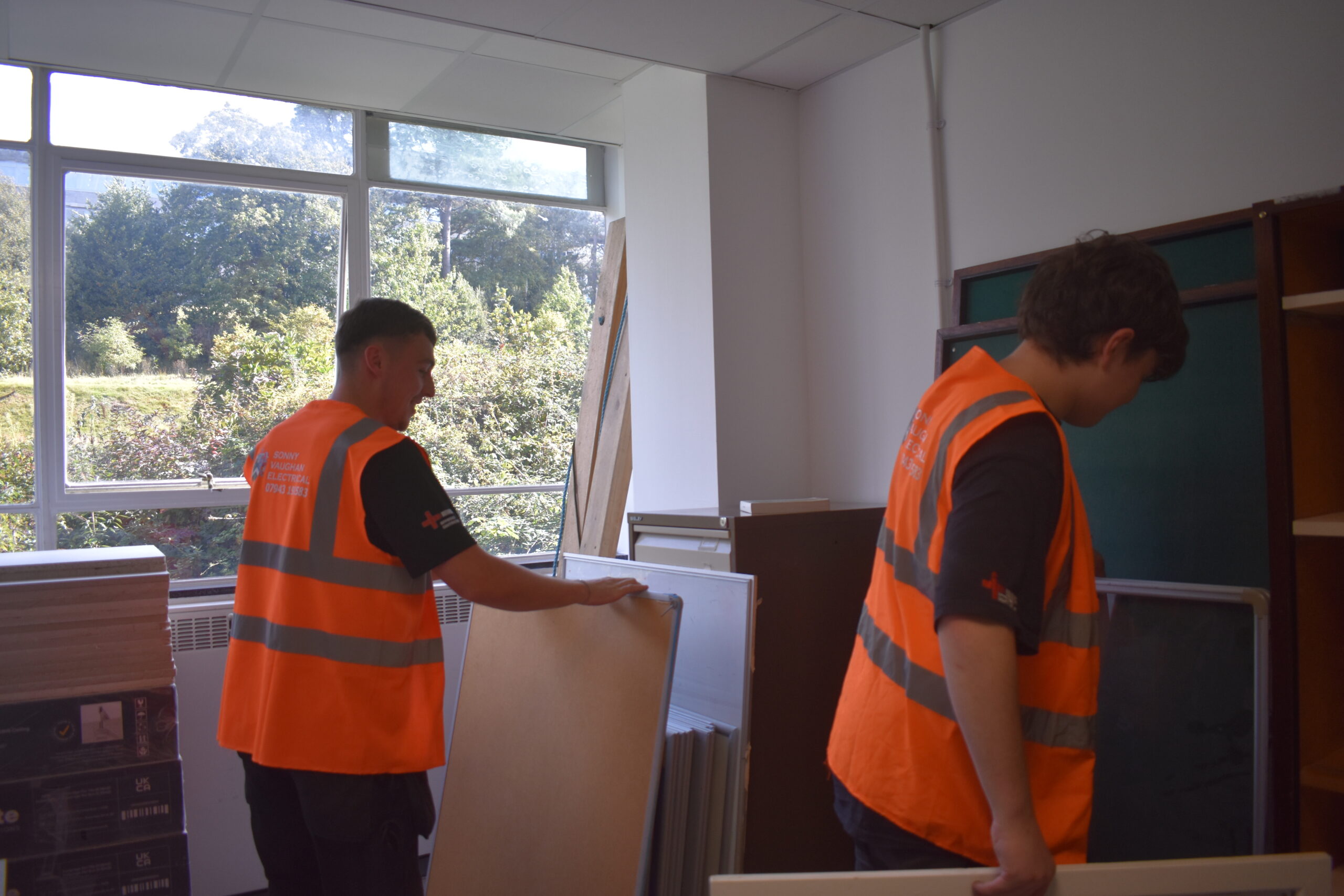 Two electricians in orange high vis jackets moving large boards in a university store room.