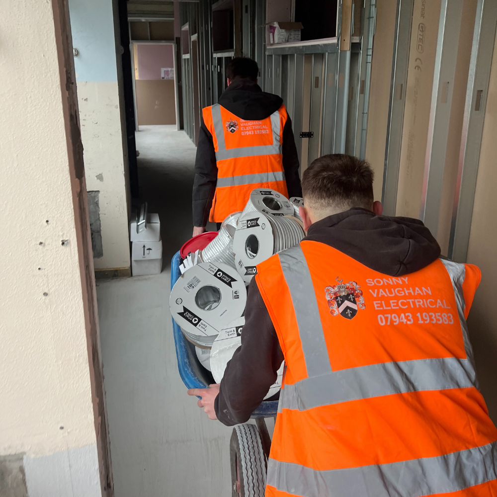 Two electricians in Sonny Vaughan Electrical orange high vis jackets. They are carrying electric wire on a commercial site.