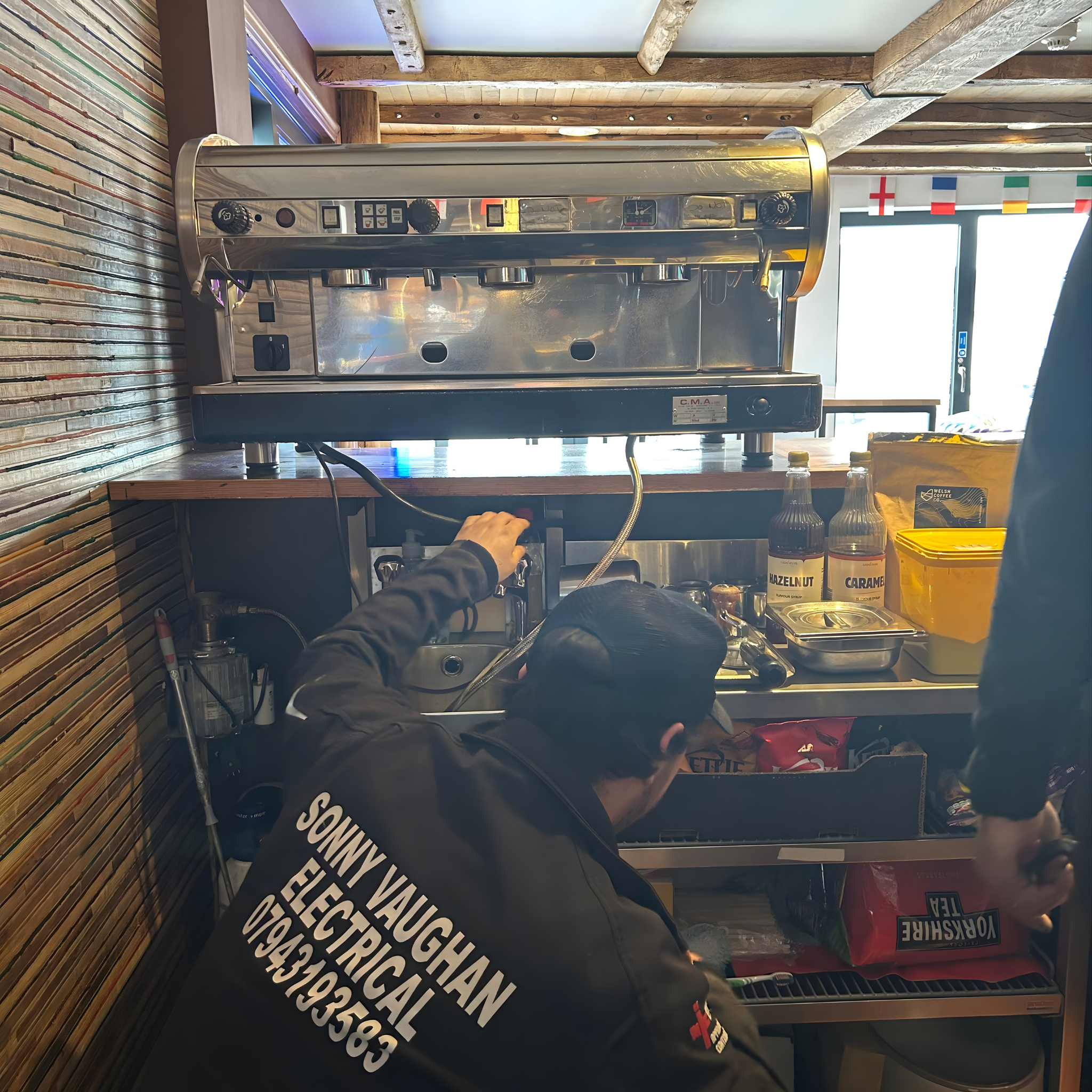 Electrician in Sonny Vaughan Electrical uniform disconnecting electric from an old metal industrial coffee machine.